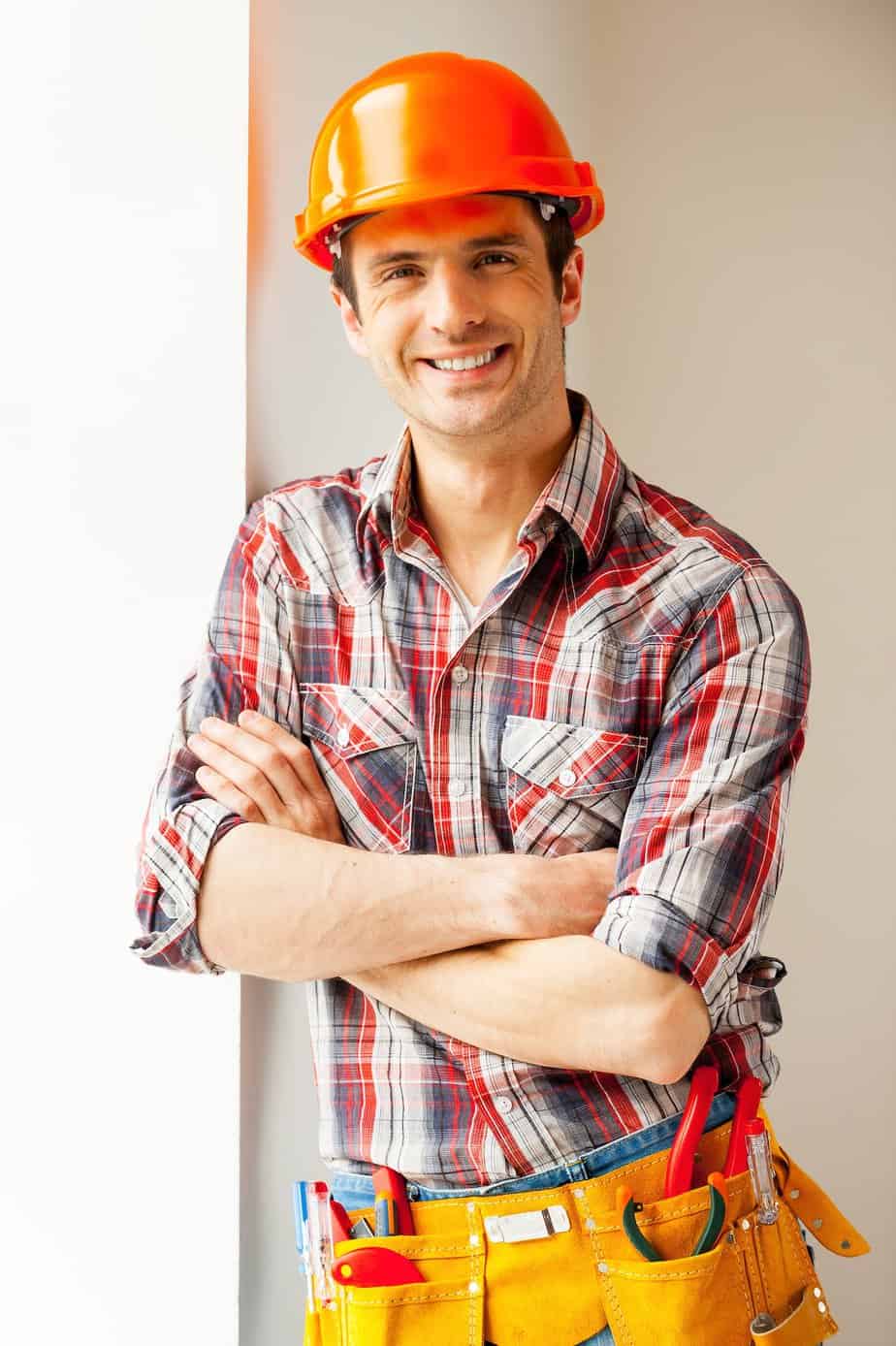 Confident repairman. Handsome young handyman in hardhat leaning at the wall and smiling at camera